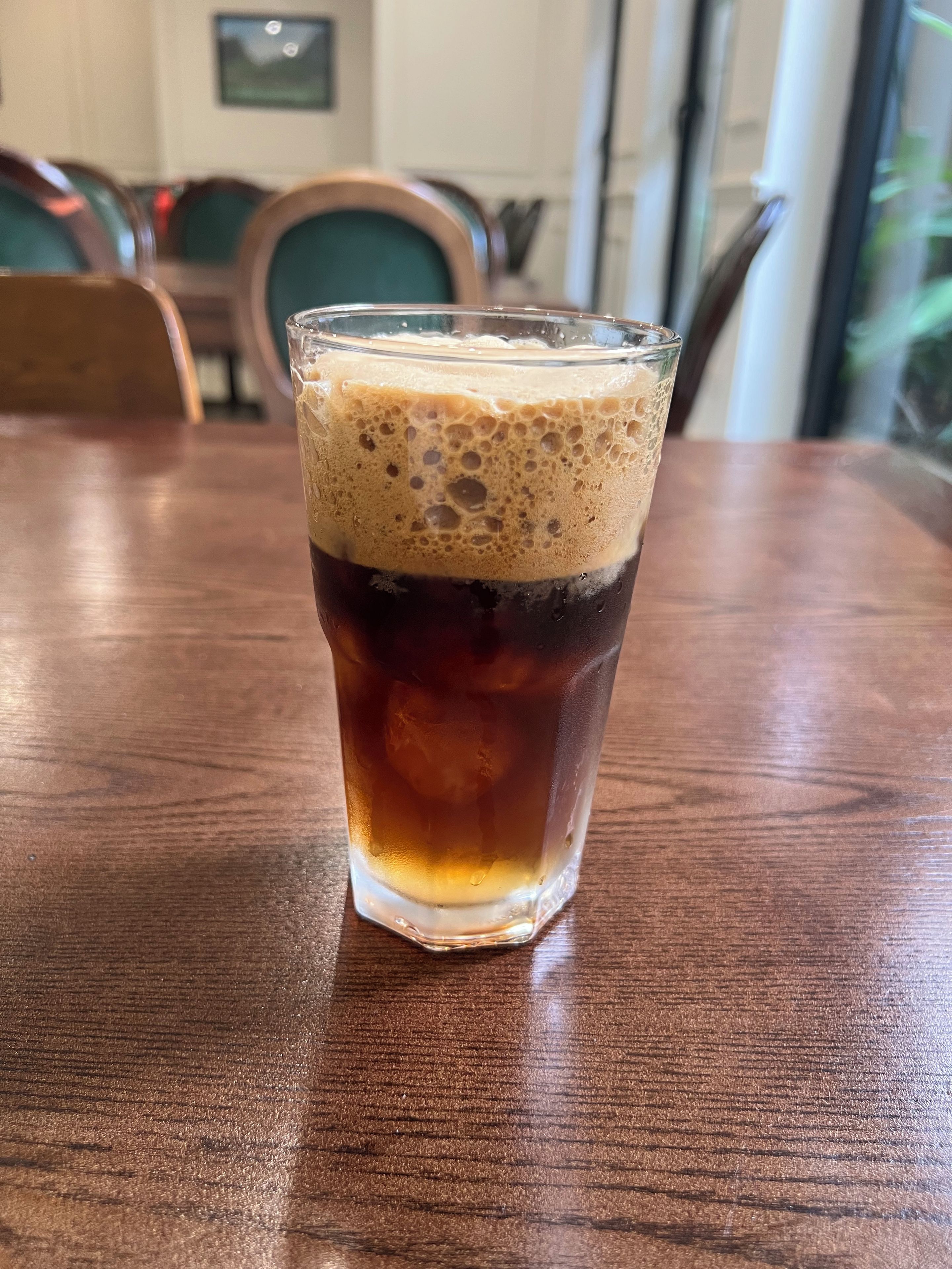 Un vaso de café helado sobre una mesa de madera en un restaurante, con sillas y una ventana al fondo.