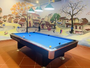 A pool table with a mural of a village scene, overhead lighting, and a billiard cue. - Paragon Noi Bai Hotel and Pool (Hanoi)