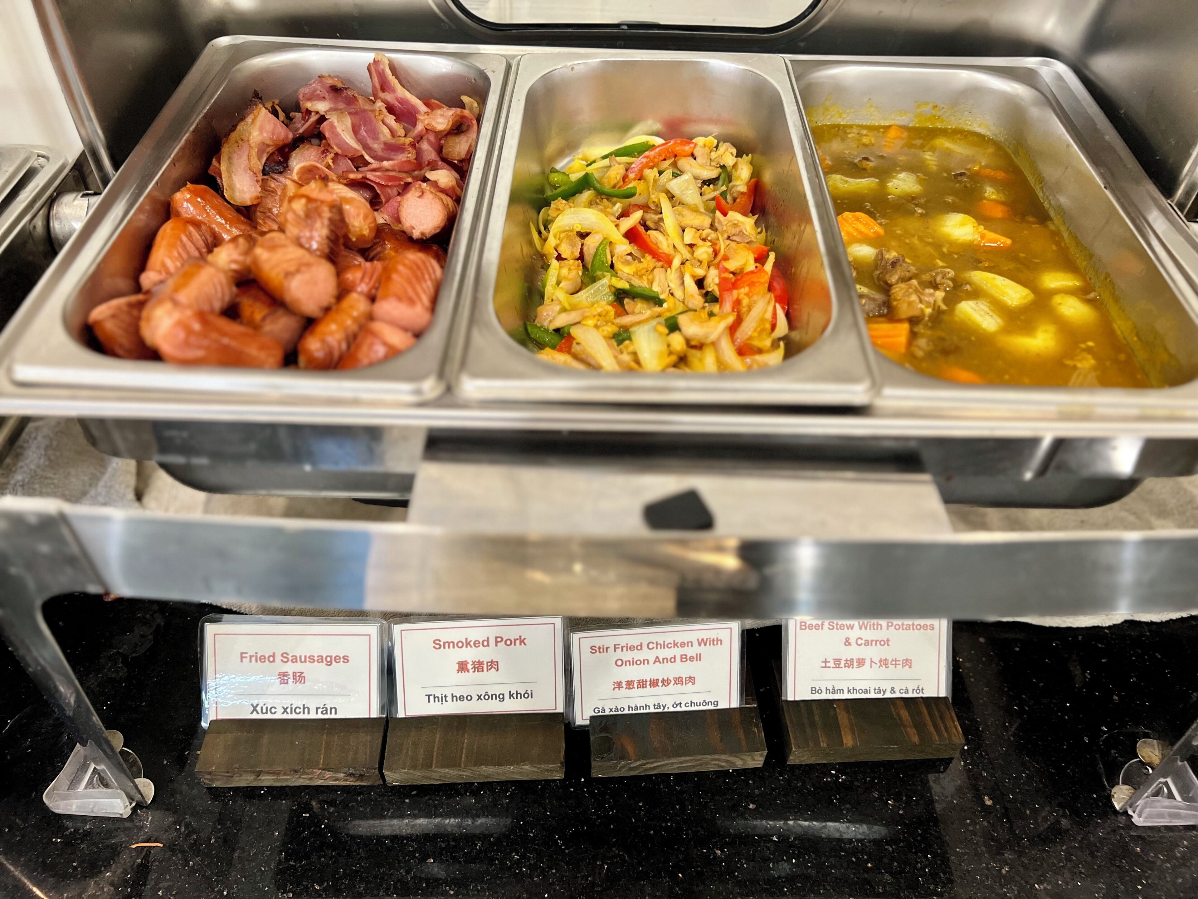 A buffet with labeled food options including fried sausages, smoked pork, stir fried chicken with onion and bell, and beef stew with potatoes and carrots.