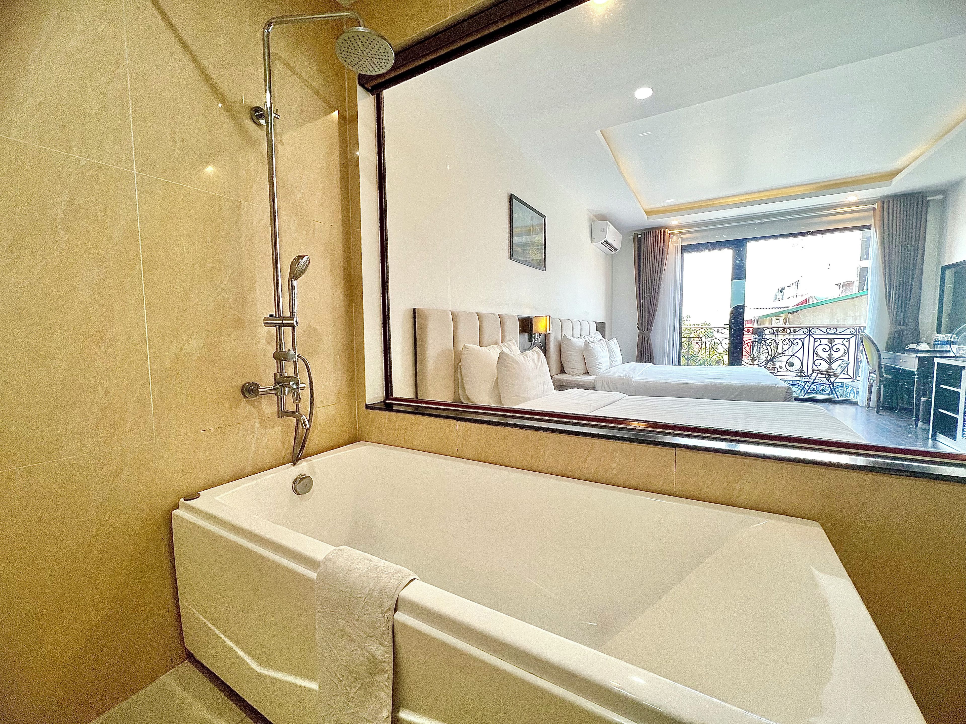 Baño de hotel con bañera, ducha y un espejo grande que refleja un dormitorio con cama y vistas al balcón.