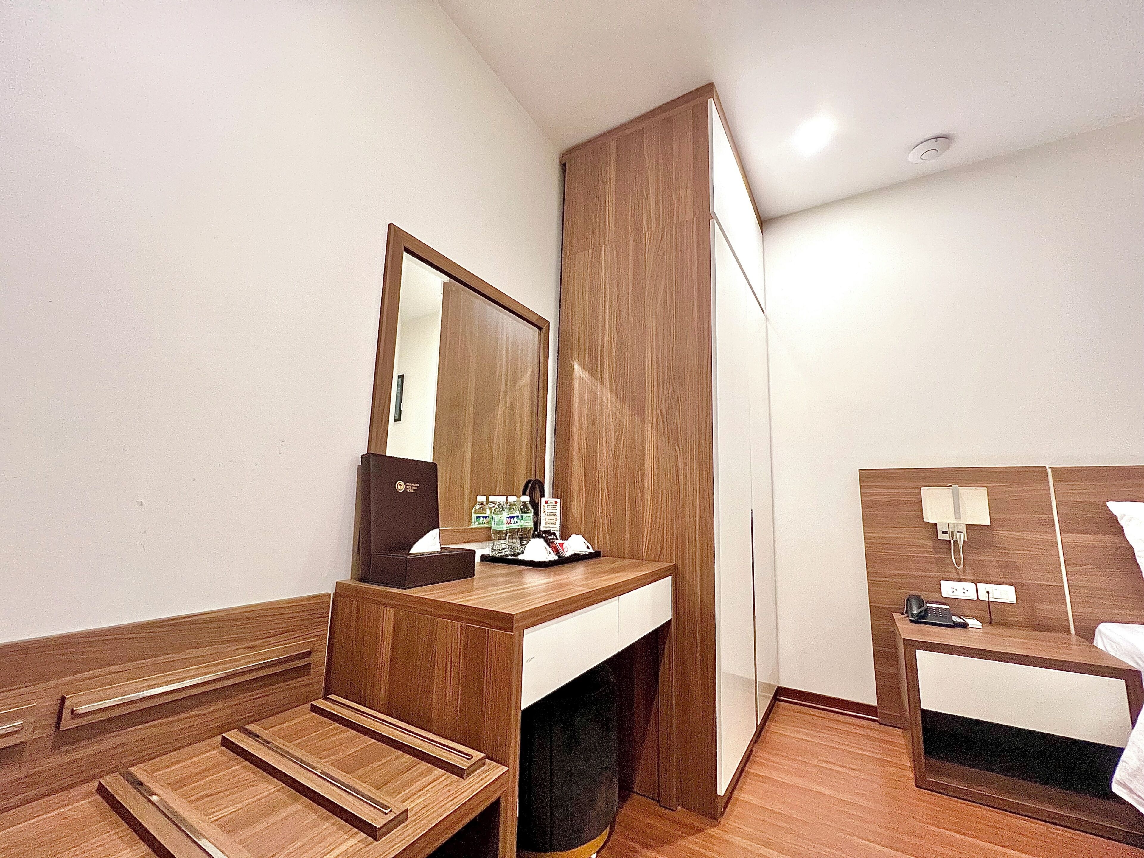 Habitación de hotel con tocador de madera, espejo, cama, mesita de noche con teléfono y pequeño escritorio.