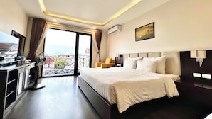 Habitación de hotel con una cama grande, un escritorio con computadora, un televisor y un balcón con vista al mar.