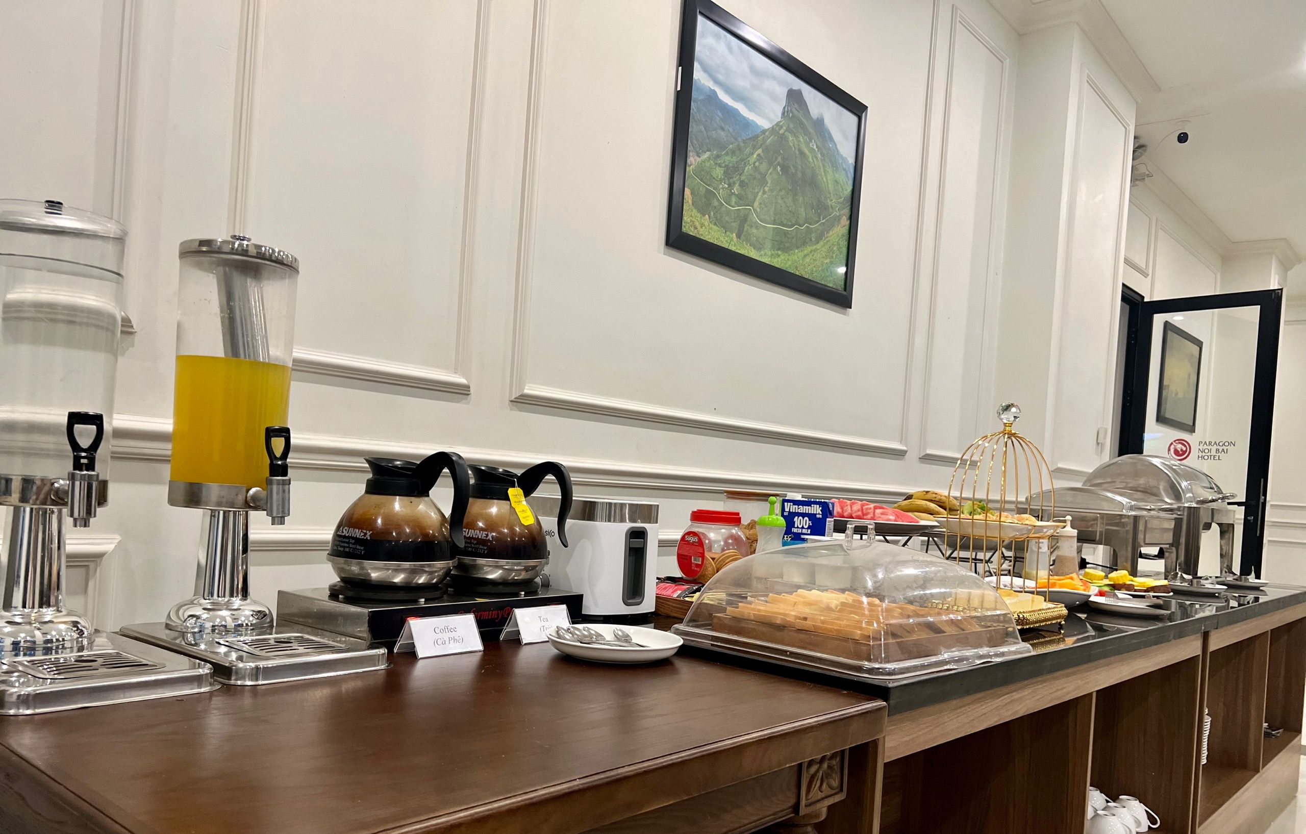 Montaje de desayuno tipo buffet con estaciones de café y té, un refrigerador Vinamilk y una variedad de alimentos.