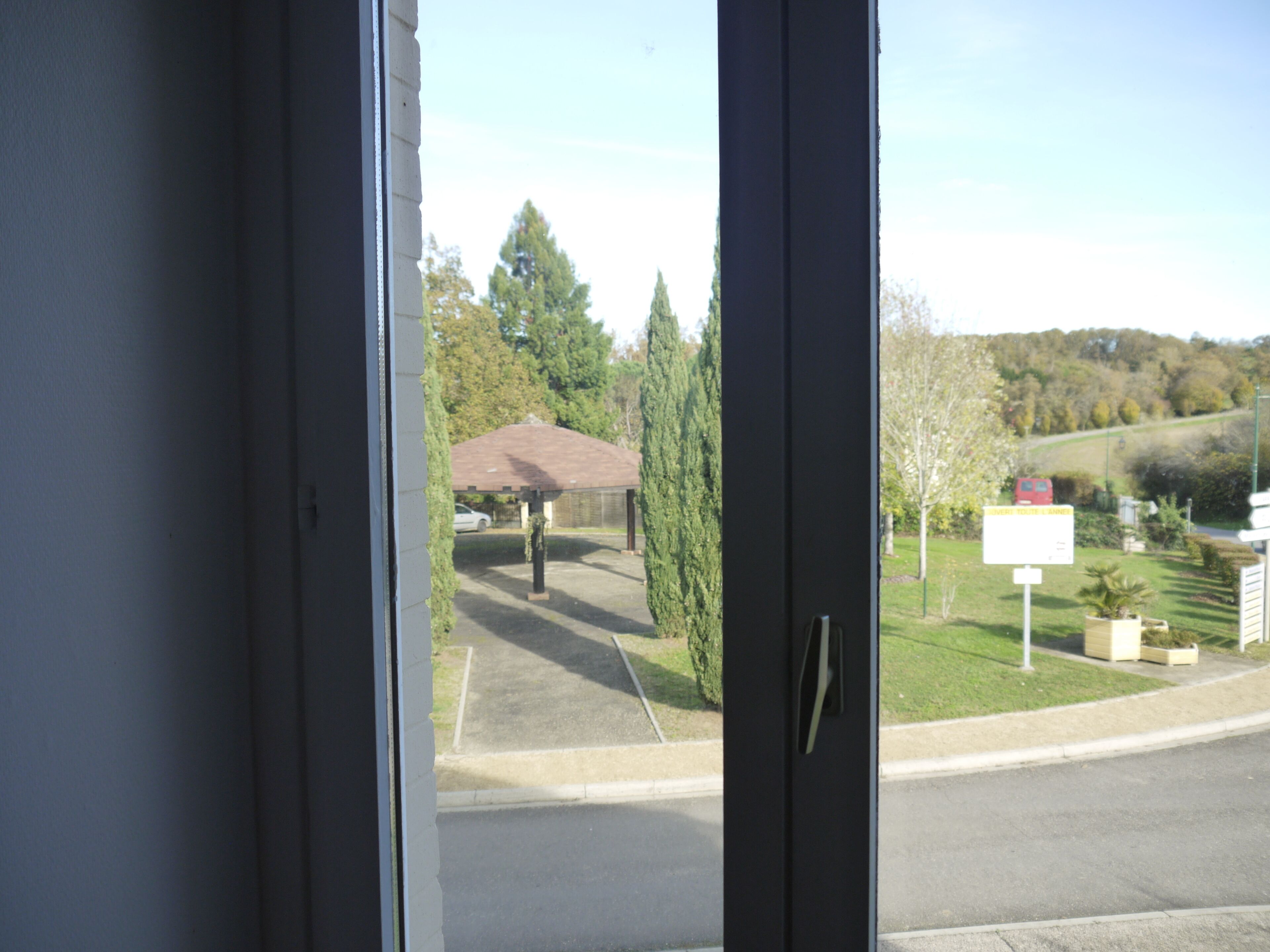 Une vue à travers une fenêtre donnant sur un parking abrité, avec un grand arbre et un panneau indicateur visible au loin.