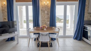 Un coin repas avec une table en bois, des chaises blanches et un saladier rempli de fruits. Un téléviseur est posé sur un meuble à gauche, et un mur en pierre est décoré d’un attrape-rêves.