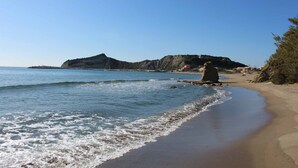 Une plage à l’eau d’un bleu limpide, bordée d’un rivage sablonneux, avec une colline rocheuse en arrière-plan.