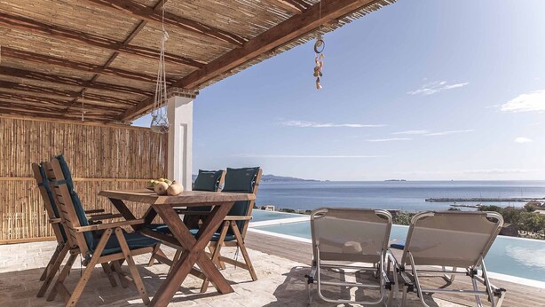 Une table et des chaises en bois, placées sous un toit de chaume, avec vue sur une piscine et la mer.