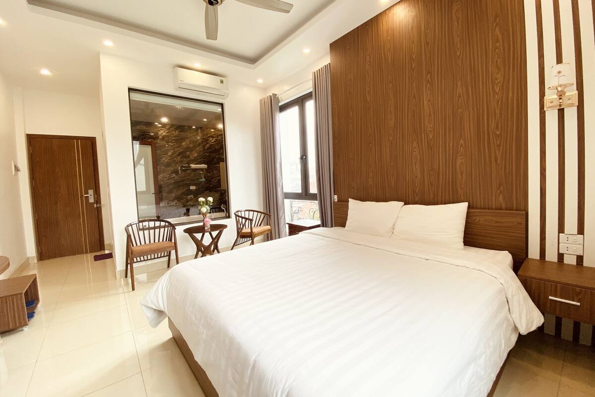 A modern hotel room with a large bed, wooden paneling, a ceiling fan, an air conditioning unit, a dining area with chairs, and a window with curtains.
