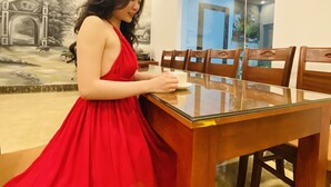 A woman in a red dress seated at a wooden dining table in a room with a mural on the wall and an air conditioning unit above. - O Zoli Home (Ninh Binh)