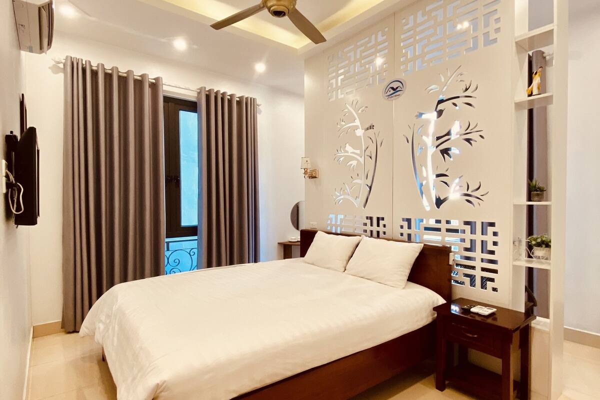 A modern hotel room with a large bed, a ceiling fan, a decorative wall panel, and a small bedside table.