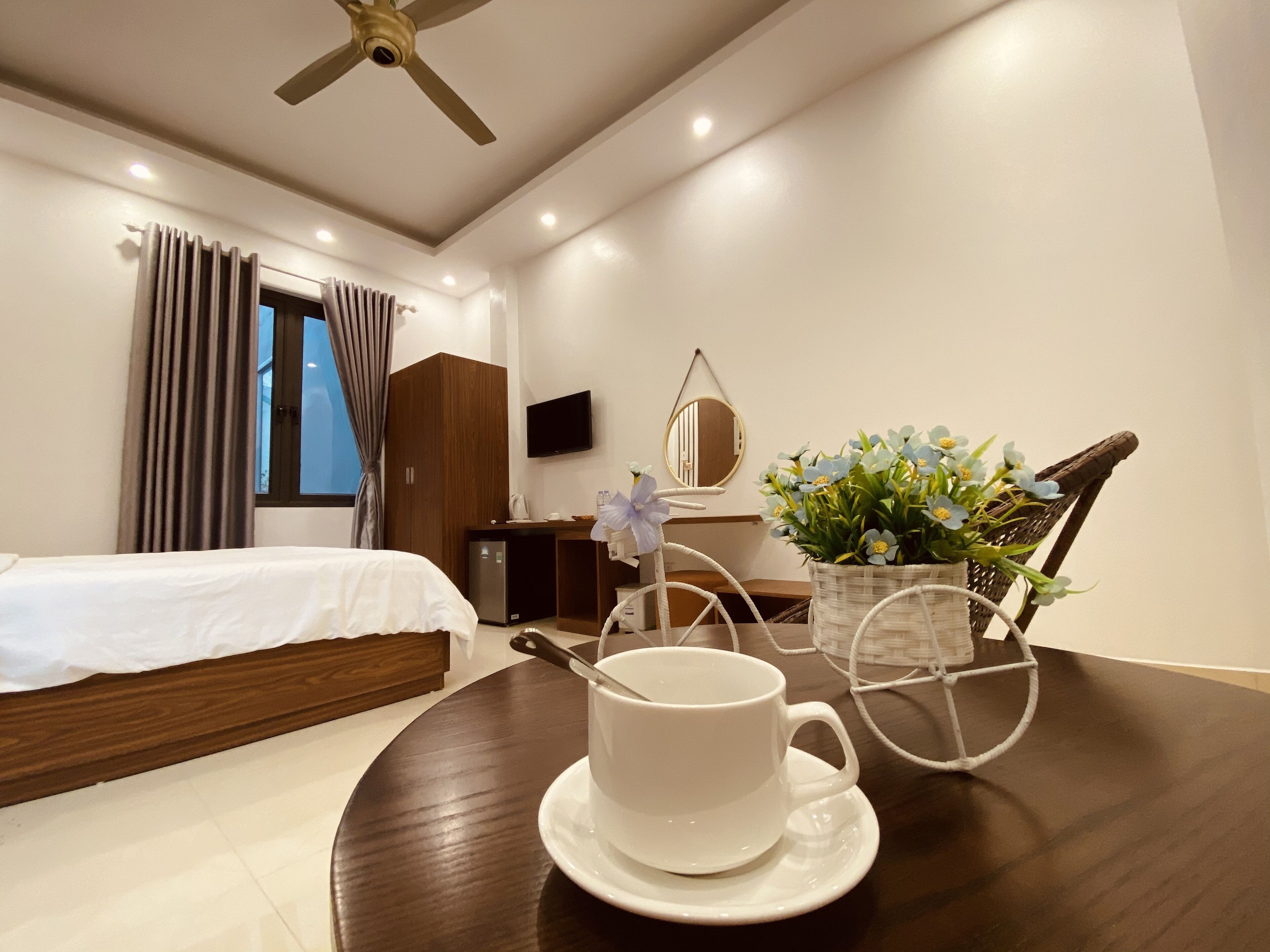 Habitación de hotel con cama, una mesa de comedor con taza y platillo, un refrigerador pequeño, televisor, espejo y ventana con cortinas.