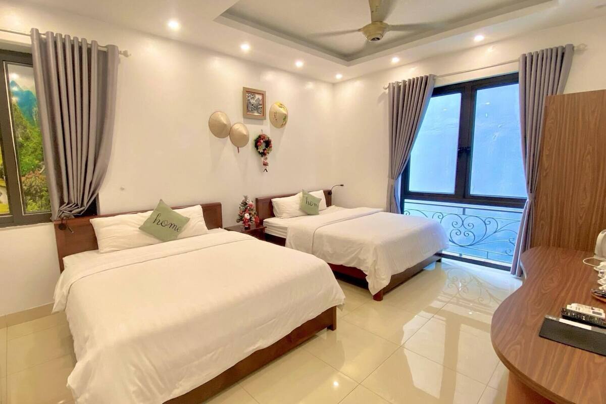 A hotel room with two beds, a large window with curtains, a small table with a lamp, and a decorative wall piece.