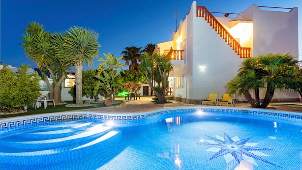 Una piscina con un diseño geométrico de azulejos, rodeada de palmeras y un edificio blanco con una escalera.