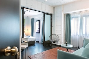 Habitación de hotel con cama, zona de estar con una silla y una mesa, y vistas al exterior a través de cortinas.