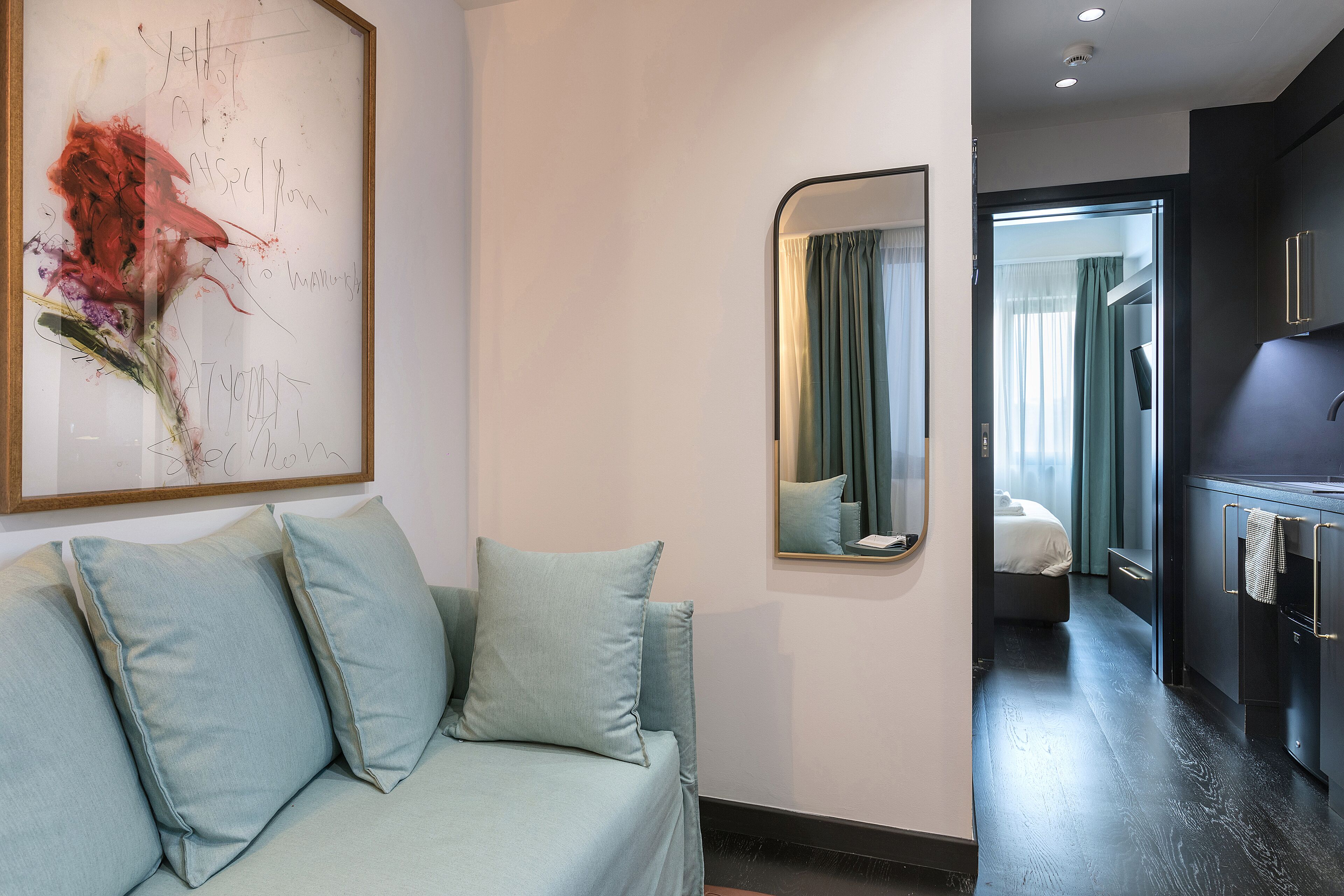 Una habitación de hotel moderna con un sofá azul claro, un cuadro abstracto grande, un espejo y vistas al dormitorio contiguo.