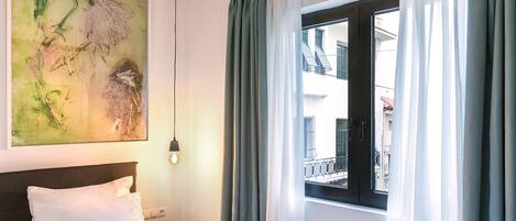 A hotel room with a bed, a nightstand, a wall-mounted lamp, a window with curtains, and a large abstract painting.