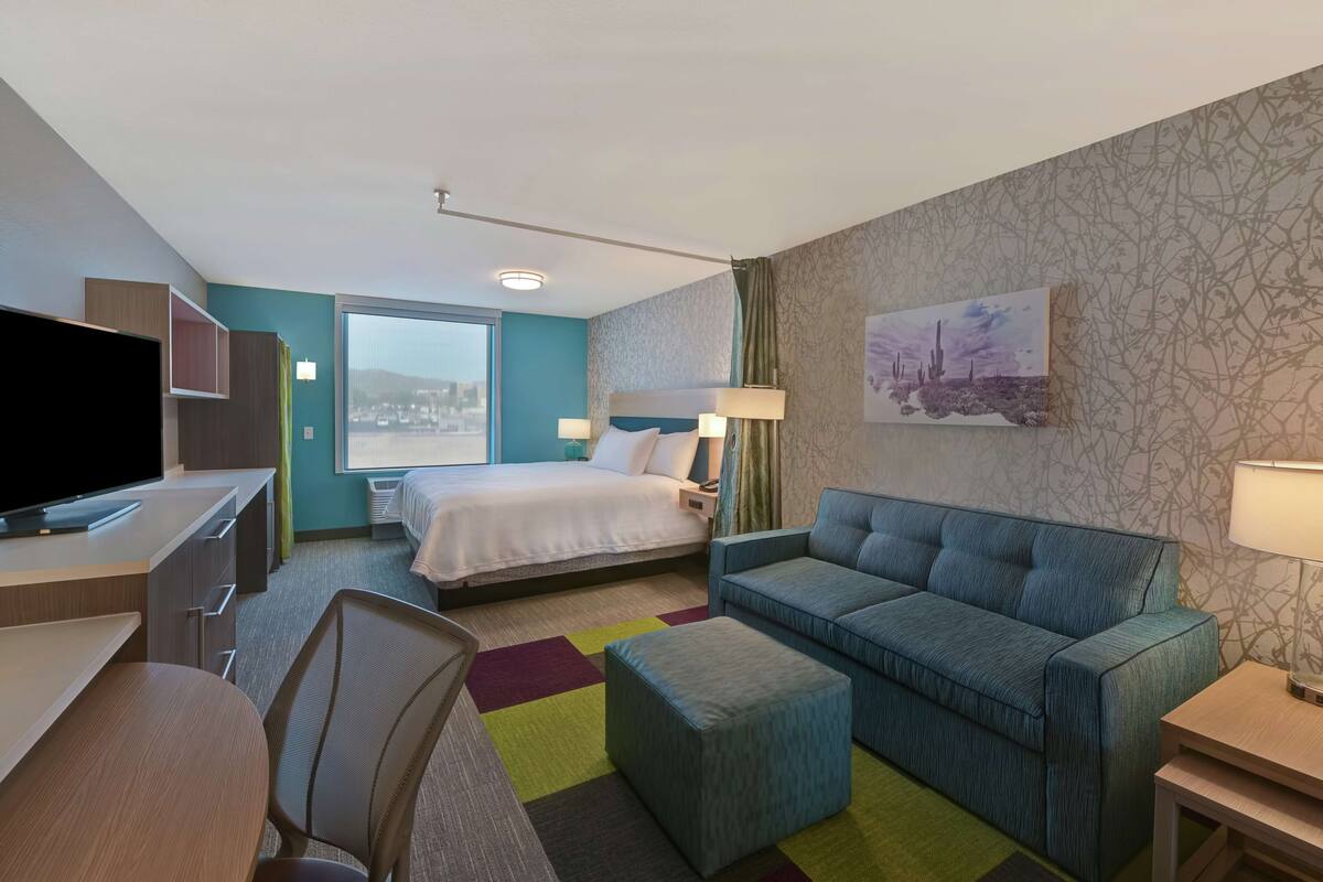 A hotel room with a bed, a sofa, a desk with a TV, and a window with a city view.