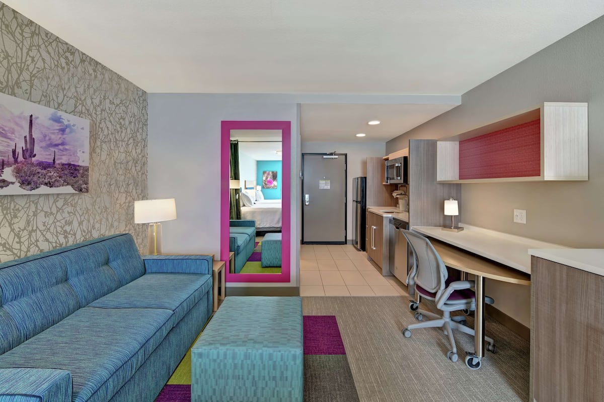 A hotel room with a sofa, a desk, a chair, a bed, and a kitchen area.
