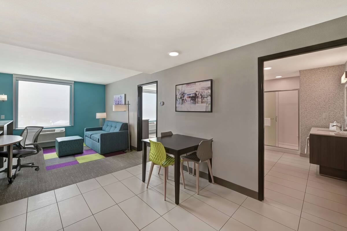 A modern hotel room with a dining area, a sofa, a table with chairs, and a bathroom visible through an open door.