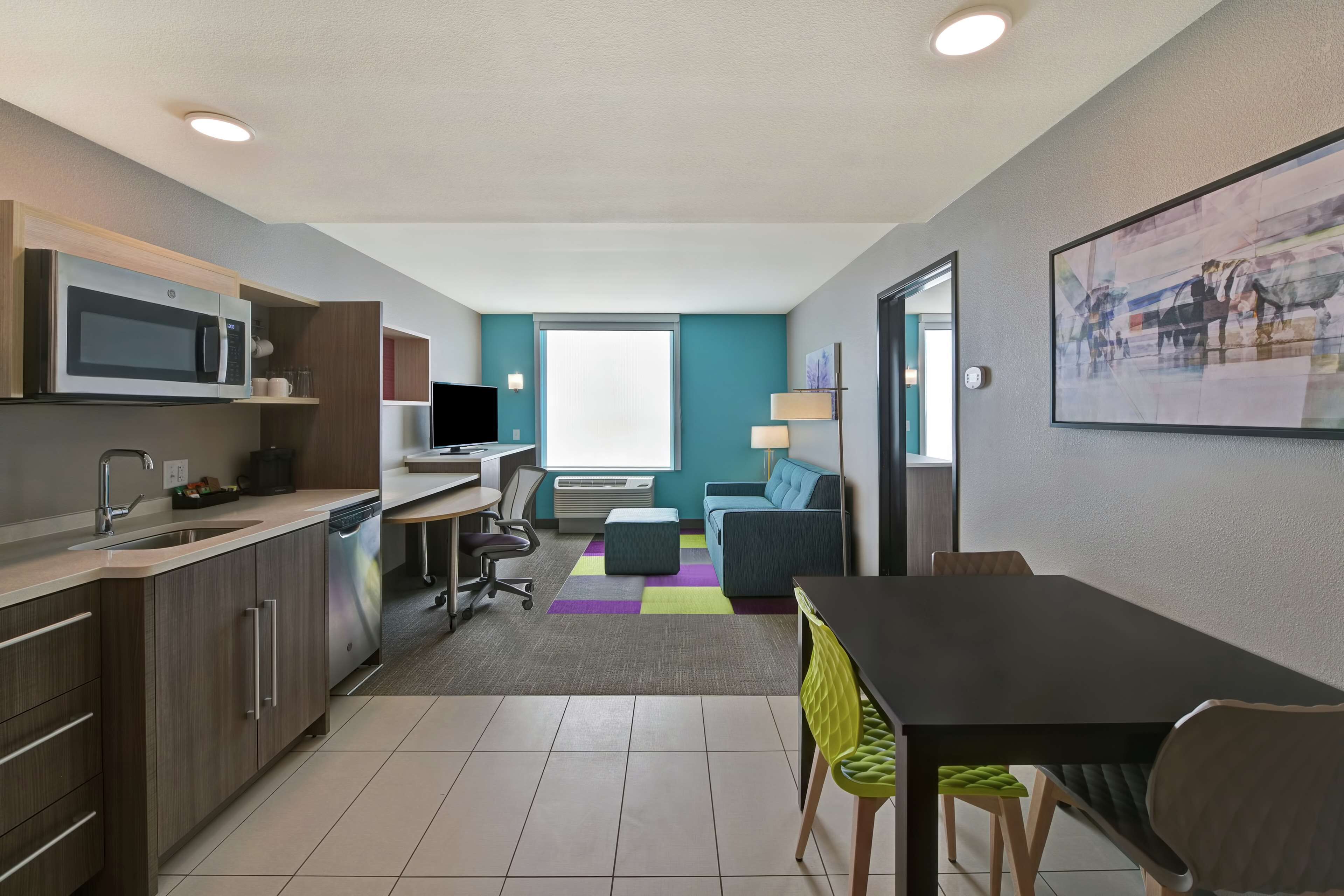 Una cocina moderna con microondas, pileta y lavavajillas. Hay una zona de comedor con una mesa y sillas. La habitación tiene un ventanal grande y un tapete colorido.