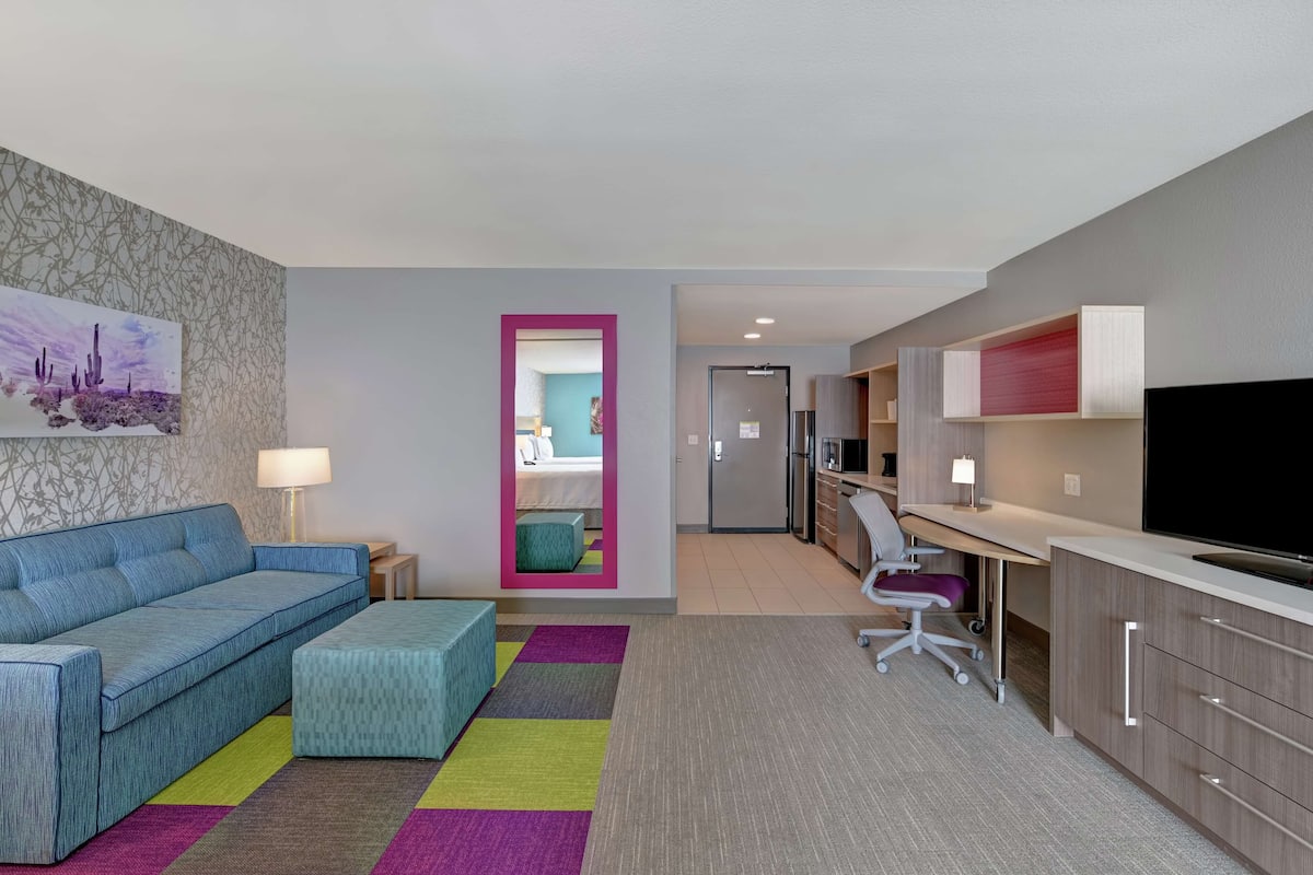 A modern hotel room with a sofa, a colorful rug, a desk with a chair, a TV, and a kitchenette.