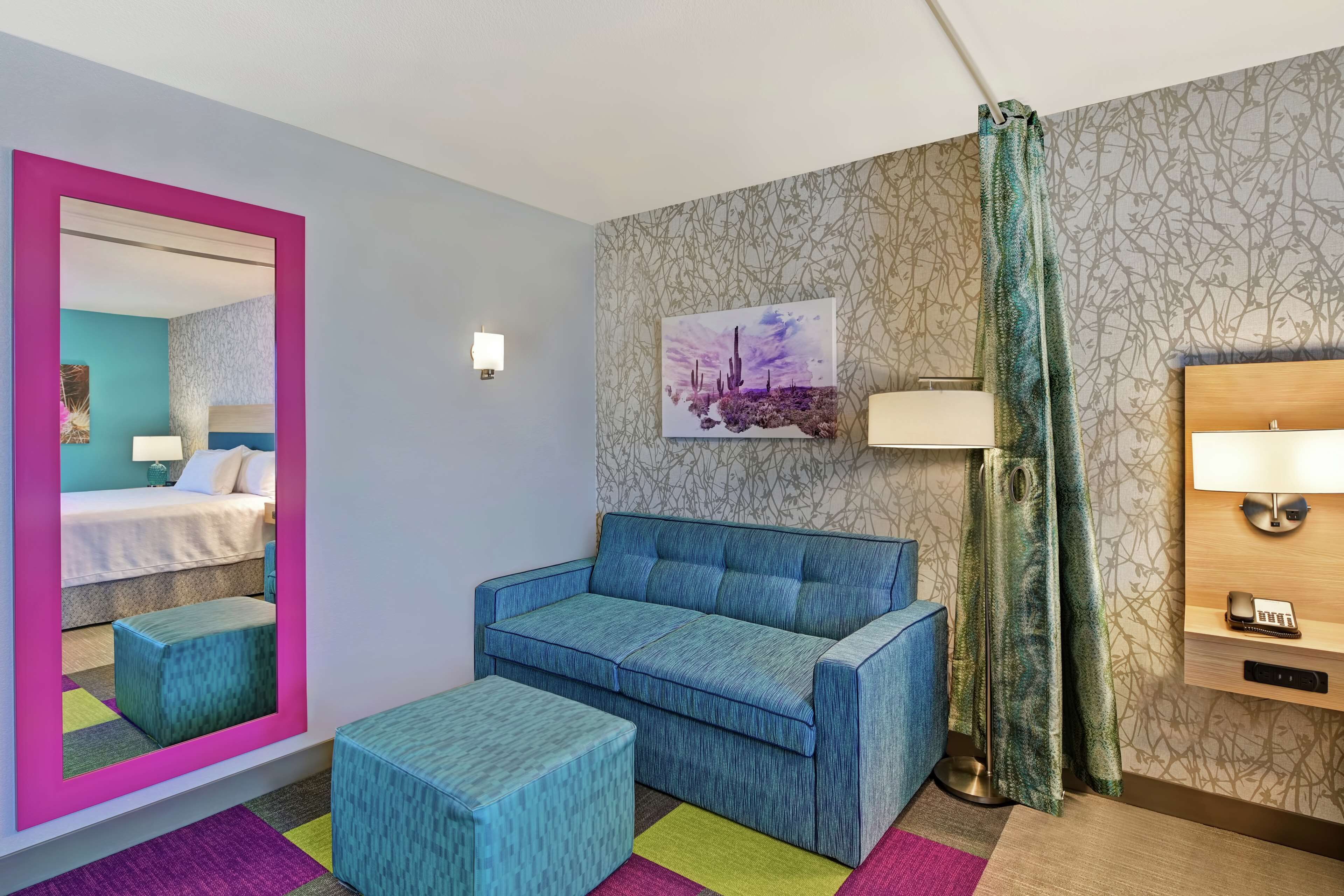 Camera d'albergo con un letto, una poltrona blu, un poggiapiedi abbinato, uno specchio con cornice rosa, una lampada a parete, un quadro e una tenda decorata.