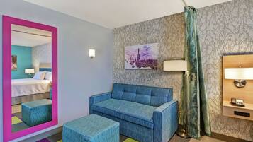 Camera d'albergo con un letto, una poltrona blu, un poggiapiedi abbinato, uno specchio con cornice rosa, una lampada a parete, un quadro e una tenda decorata.