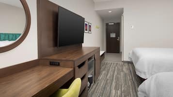A hotel room with a wooden desk, a flat-screen TV, a mirror, a bed, and a carpeted floor.