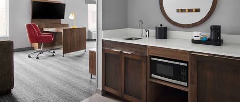 A modern bathroom with a wooden vanity, a round mirror, a sink, and a coffee maker.