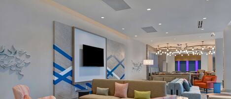 A modern hotel lobby with a sofa, striped chairs, a round wooden coffee table, and a TV mounted on the wall.