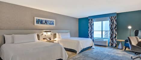 A hotel room with two beds, a desk, a chair, a window with curtains, and a wall-mounted artwork.