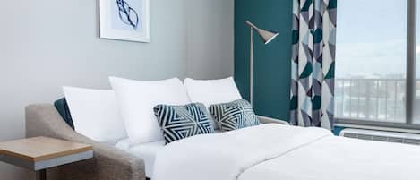 A modern hotel room with a bed, a side table, a floor lamp, and a window with curtains.