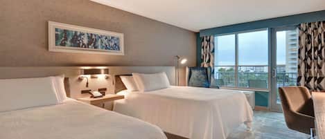 Habitación de hotel con dos camas, un escritorio, una silla, una lámpara y un ventanal con vistas.