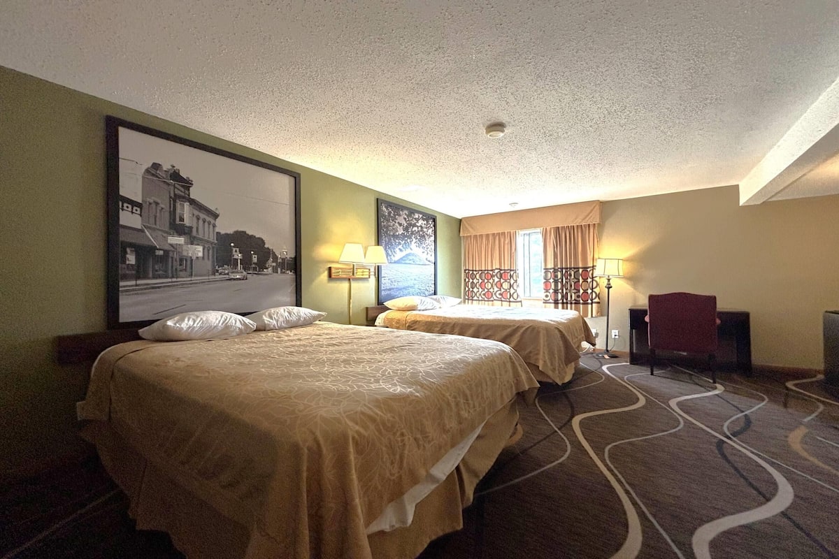 A hotel room with two beds, a desk, a chair, a lamp, and a framed picture on the wall.