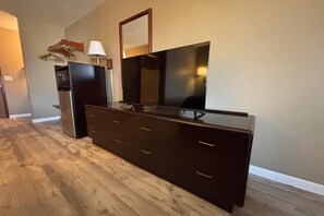 Un aparador de madera oscura con un televisor de pantalla plana, una lámpara y un perchero en una habitación con piso de madera y paredes color beige.