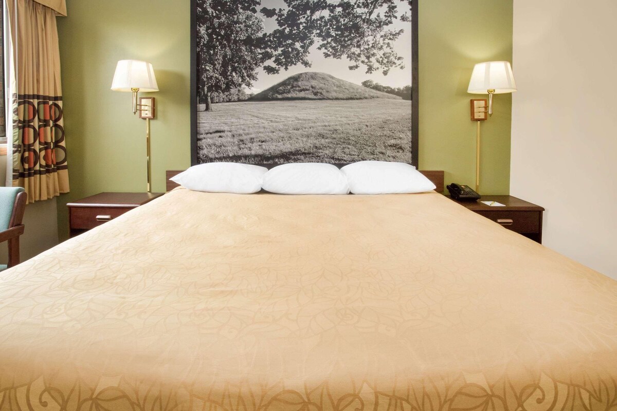 Habitación de hotel con una cama grande, dos mesitas de noche con lámparas, una mesita auxiliar y un cuadro grande en la pared.