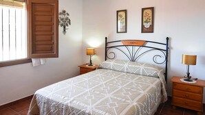 A bedroom with a bed, two nightstands, a window with blinds, and wall art. - Casa Carrerilla (Frigiliana)