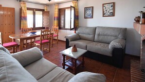 A living room with a sofa, a dining table with chairs, and a kitchen area with a stove. - Casa Carrerilla (Frigiliana)