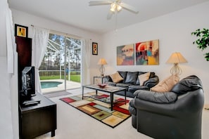 A modern living room with a black leather sofa, a coffee table, and a TV on a stand. There are two lamps, a painting, and a view of a pool through the glass door.