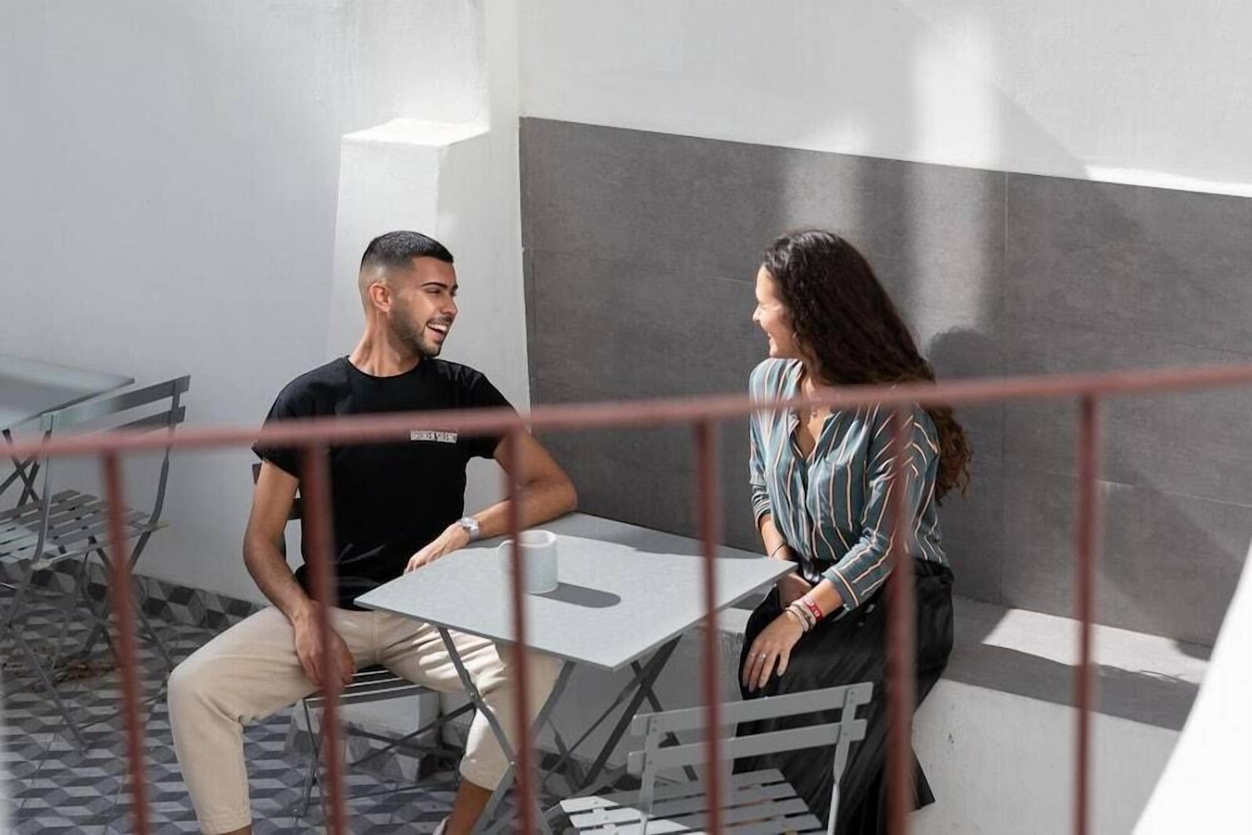 Two people sitting at a table in a modern indoor space with a geometric floor pattern and a large grey wall.