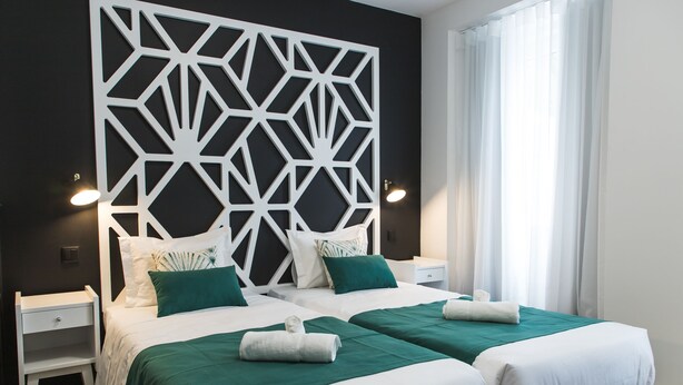 Une chambre d’hôtel avec deux lits, du linge de lit blanc, des coussins décoratifs verts, une tête de lit blanche ornementale et une table de chevet avec une lampe.