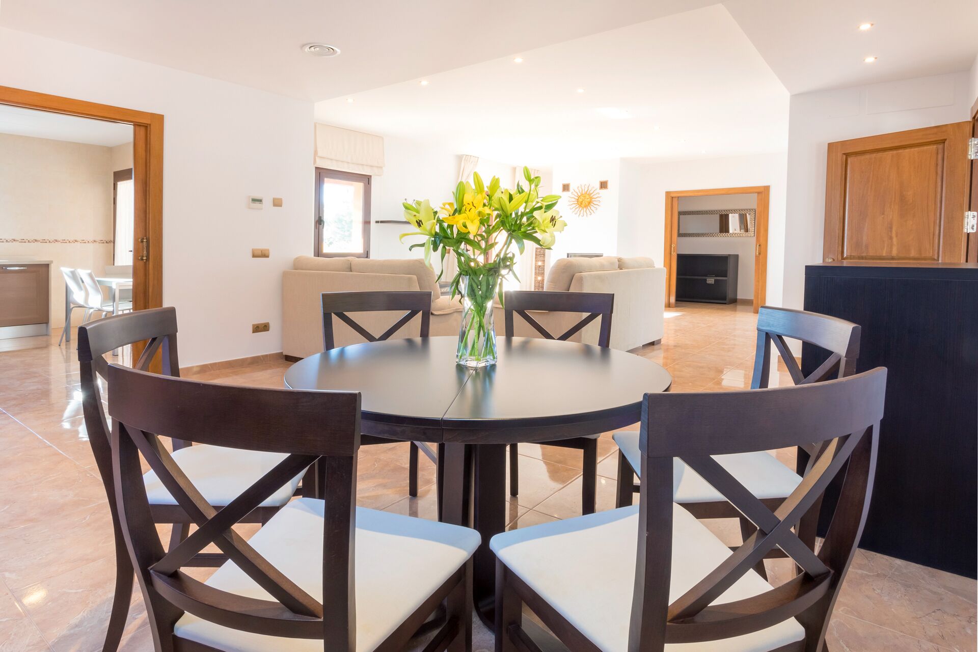 A dining area with a round table and six chairs, a vase with flowers, and a kitchen in the background.