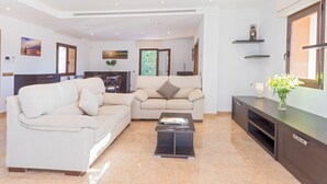 A spacious living room with a sofa, a coffee table, and a TV stand. There is a dining area in the background with a table and chairs. - Villa Near Ibiza Town Sleeps 12/14 (Cap Martinet)