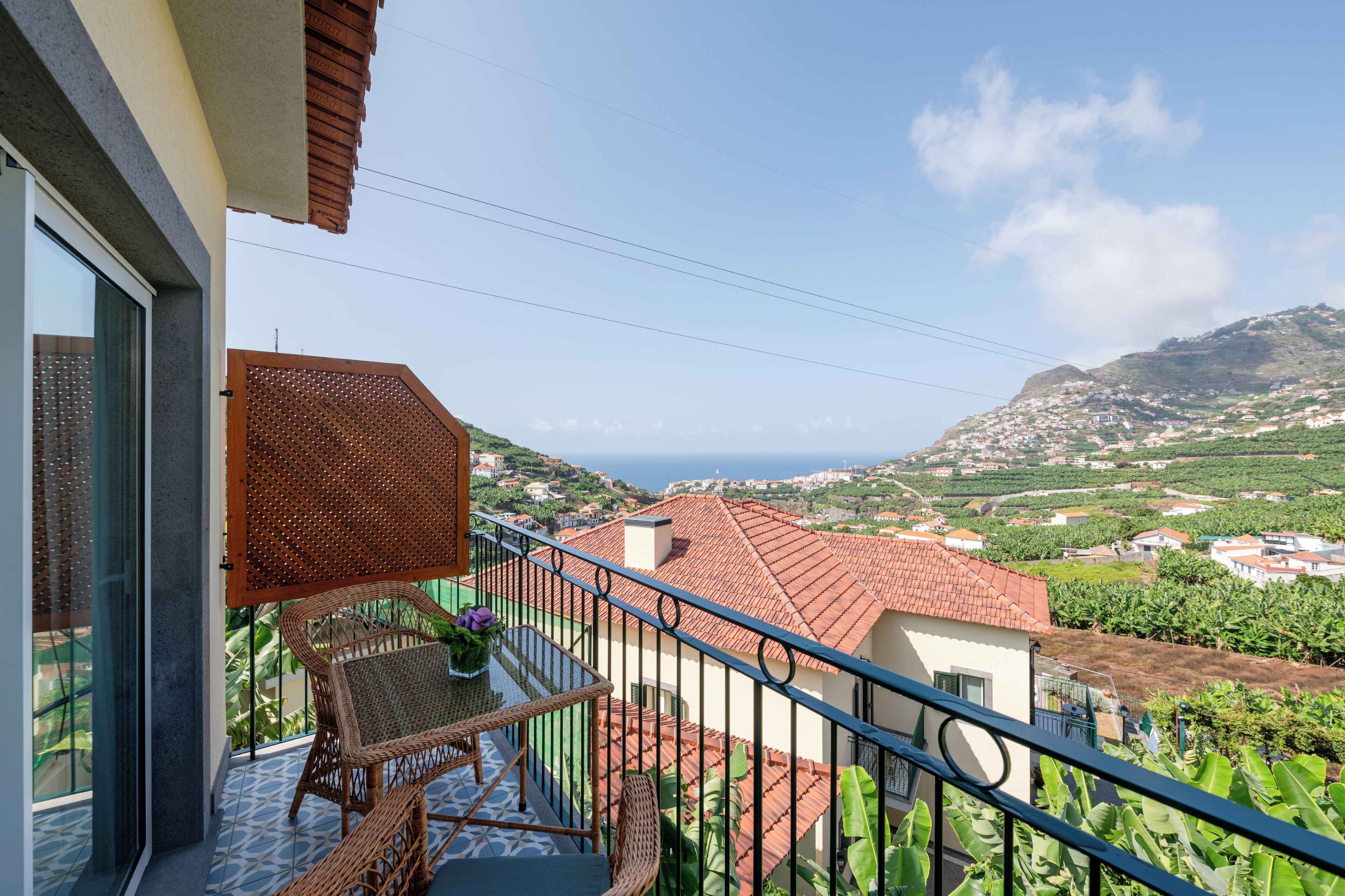 A balcony with wicker furniture, a view of a coastal town, and a tiled floor.