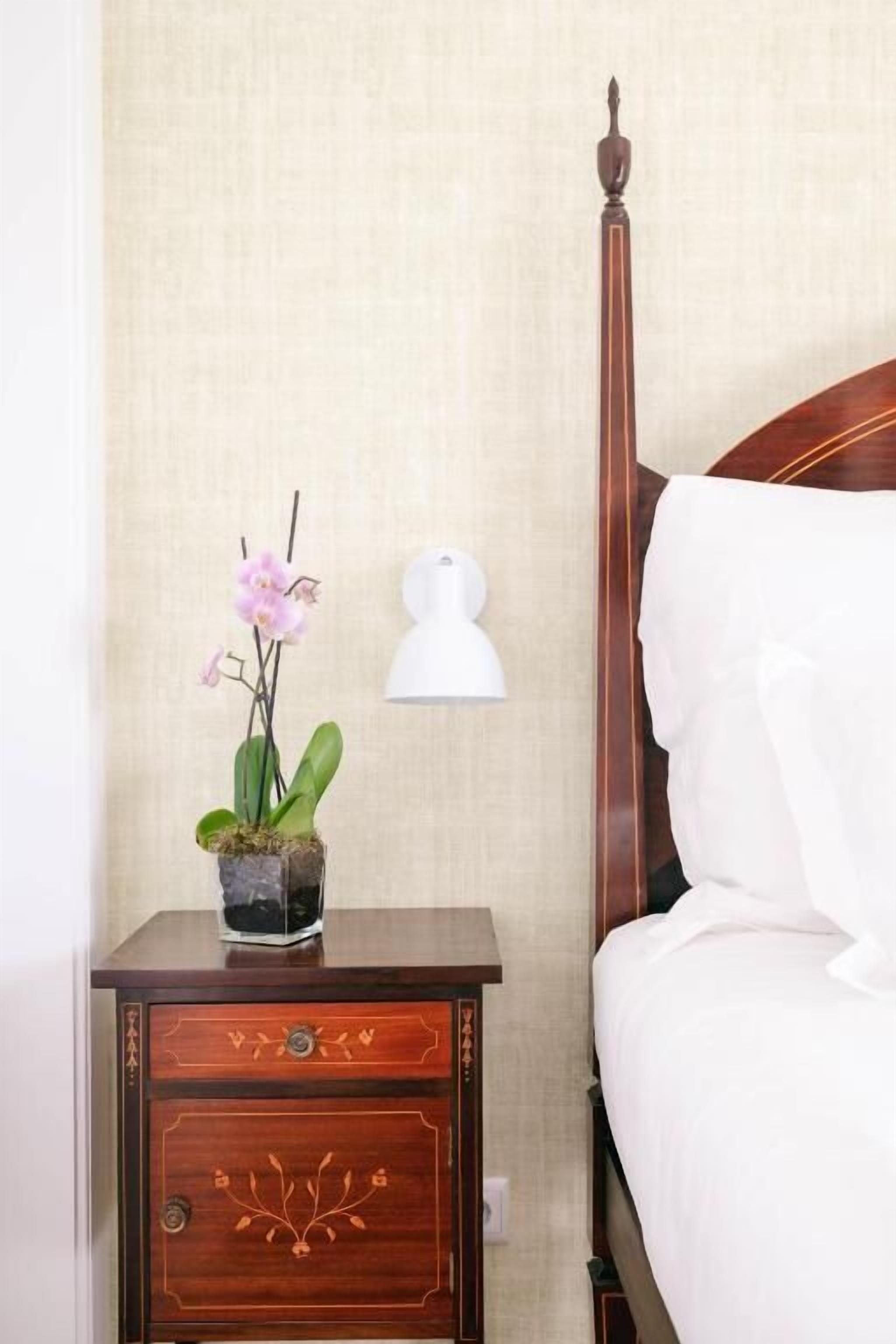 A neatly made bed with white linens, a wooden nightstand with a drawer, a potted orchid, and a wall-mounted lamp.