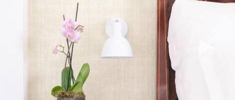 A neatly made bed with white linens, a wooden nightstand with a drawer, a potted orchid, and a wall-mounted lamp.