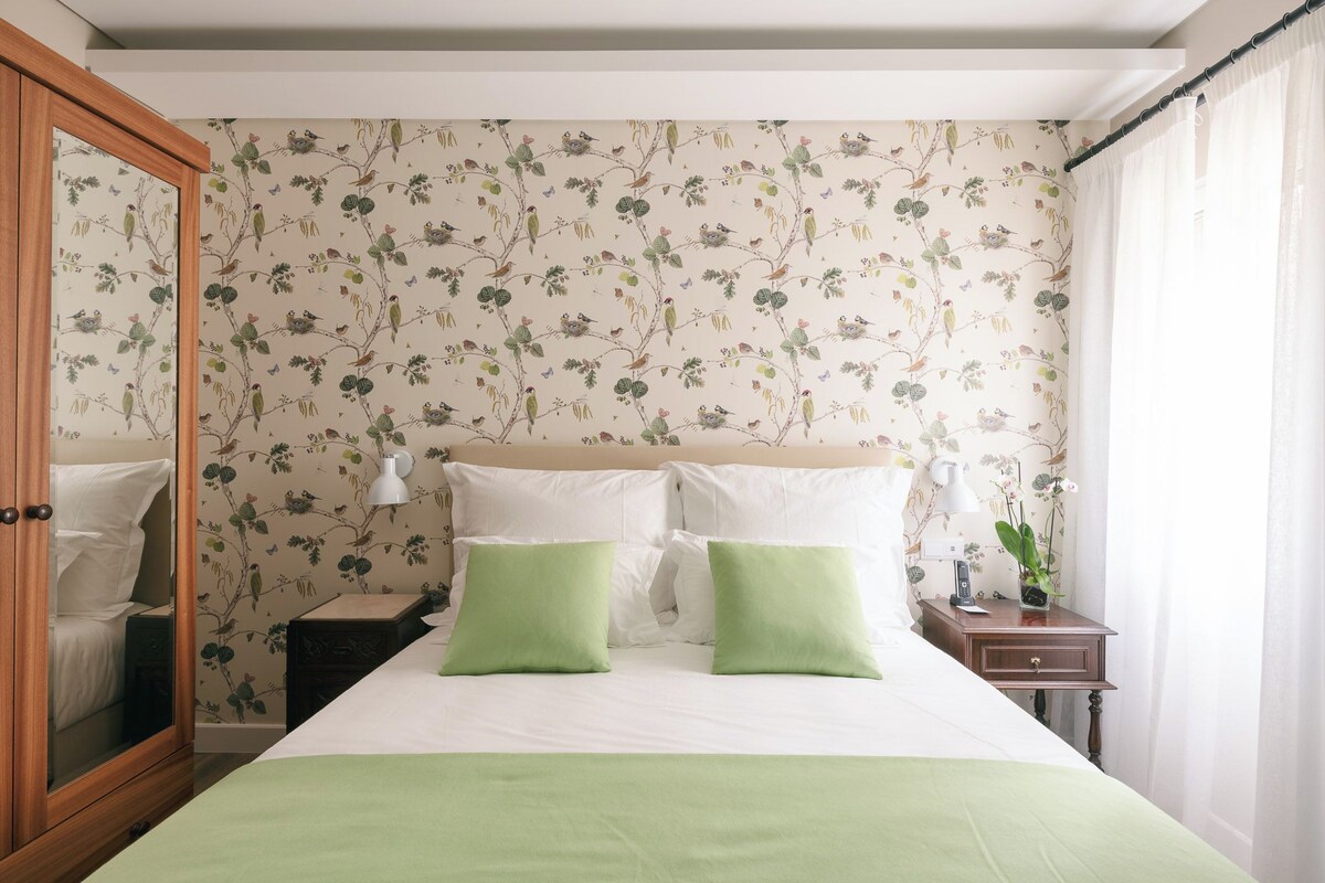 A bedroom with a bed, two green pillows, a nightstand with a lamp, and a mirror.