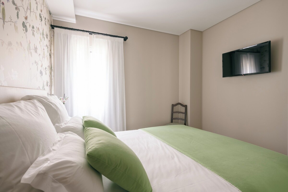 A bedroom with a bed, a window with curtains, a chair, and a flat-screen TV mounted on the wall.