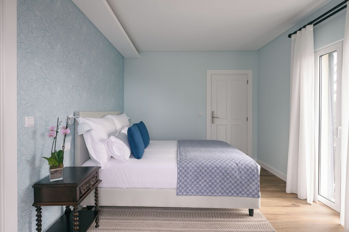 A bedroom with a bed, a nightstand, a potted plant, and a window with curtains.