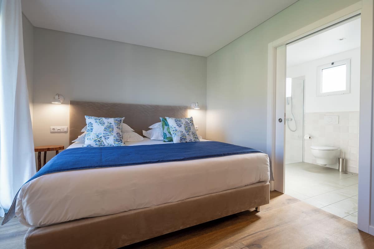 A bedroom with a bed, bedside lamps, a wooden floor, and a bathroom visible through an open door.